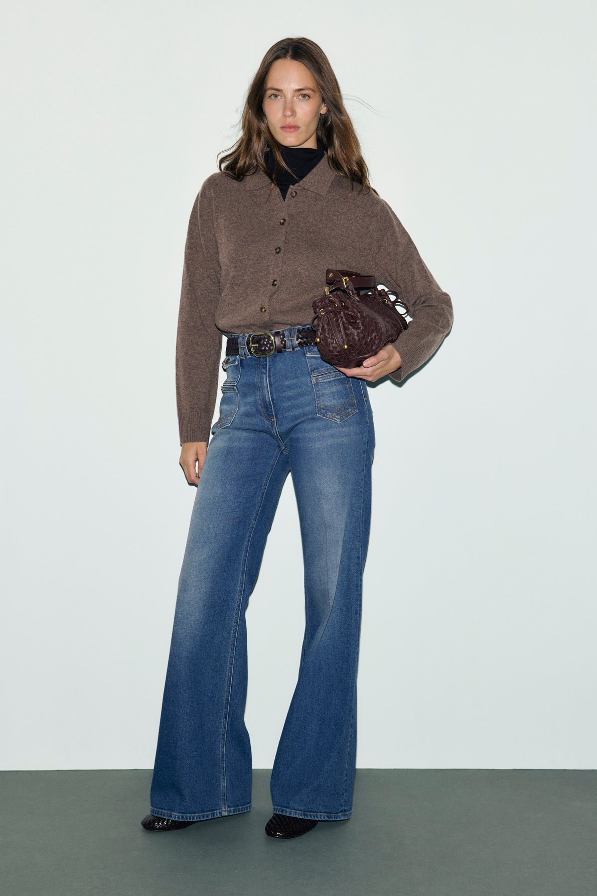 Woman stands against a plain background, wearing a brown button-up cardigan, high-waisted jean flare blue pants, a black turtleneck, and black shoes. She holds a small dark brown bag at her hip.