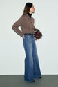 Woman standing sideways, wearing a brown sweater over a black turtleneck, and blue pantalon évasé jeans with a taille haute fit, holding a brown handbag. She has long hair and is looking forward against a plain light background.