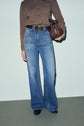 A person wearing a brown long-sleeve top, high-waisted blue jean flare pants, a belt, brown shoes, and holding a small brown purse stands against a plain background.