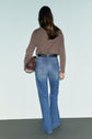 A woman with long brown hair stands facing away, wearing a brown sweater and blue high-waisted jeans à taille haute with a slight jean flare. She holds a textured handbag in her left hand, while the background features soft shades of light blue and gray.