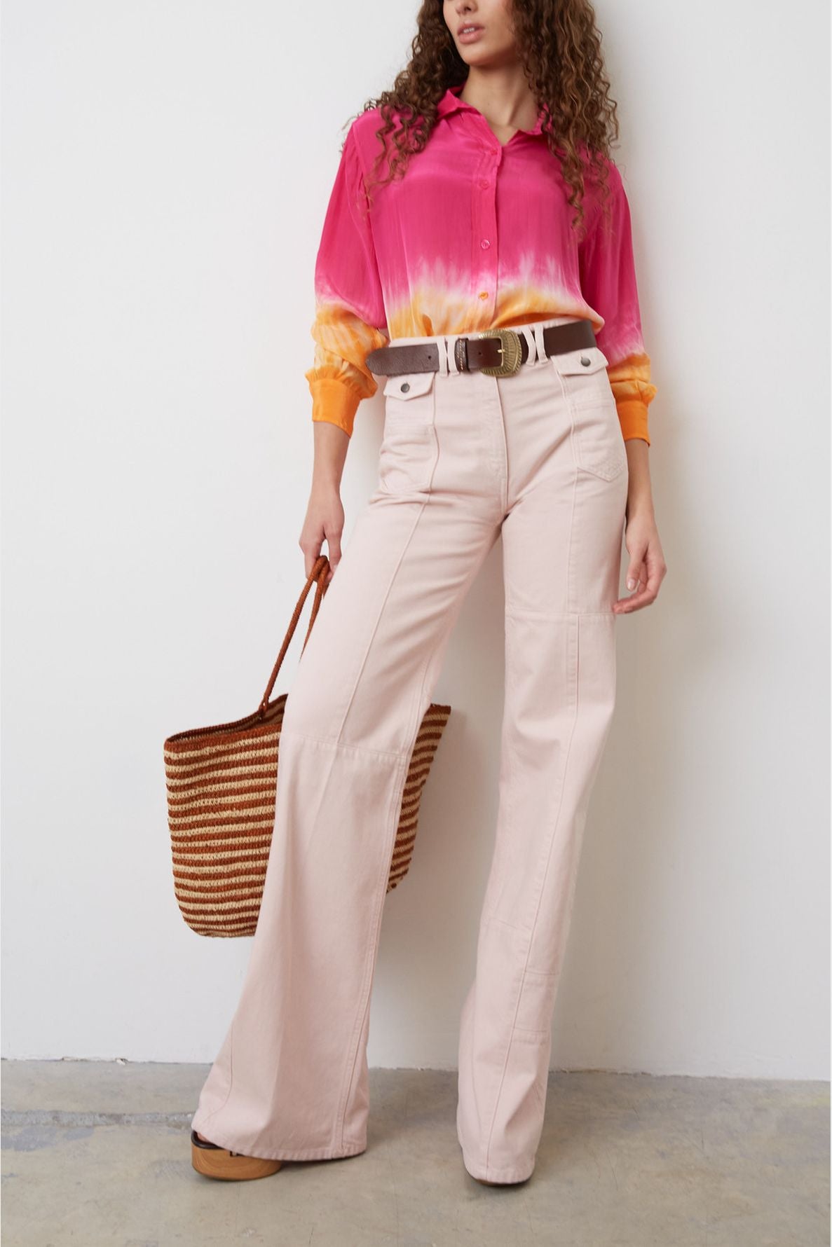 A woman wearing a pink and orange tie-dye shirt, wide-leg pale pink pants, brown belt, and holding a striped woven tote bag stands against a white wall.