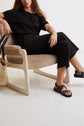 A woman with long curly hair sits in a beige chair, wearing a black top, black pants, and black sandals. A woven straw tote bag with black handles rests beside the chair on a white floor.