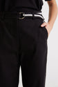 Person wearing black pants and a black shirt, with their right hand in their pocket. The pants have a black and white striped belt with a silver buckle. Only the lower torso and upper legs are visible.