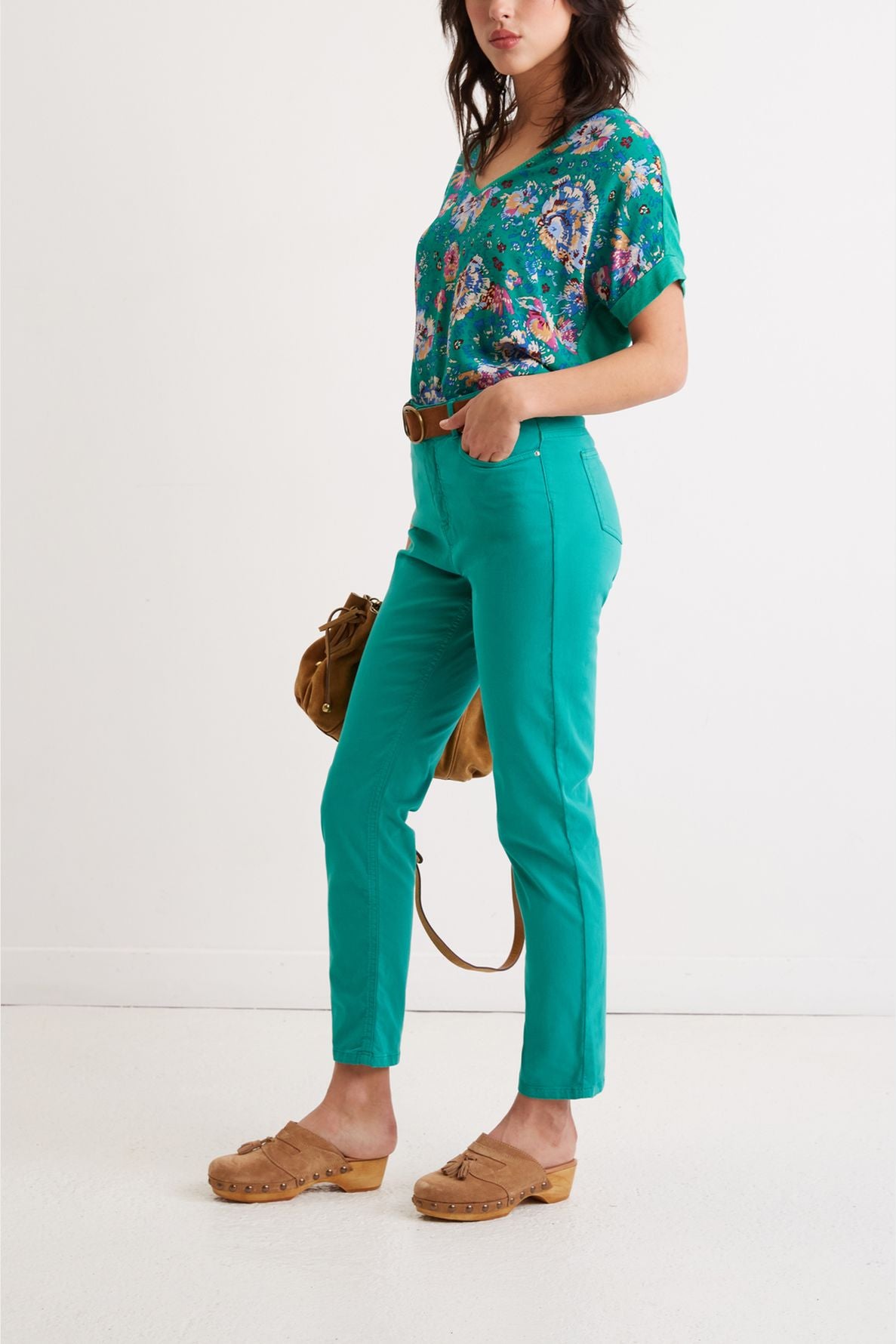 A woman stands sideways wearing a floral top, teal pants, brown belt, tan clogs, and holding a brown handbag against a plain white background.