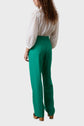 A person with long curly hair wears a white blouse with puffed sleeves and high-waisted green pantalon large, standing with their back to the camera against a plain light background.