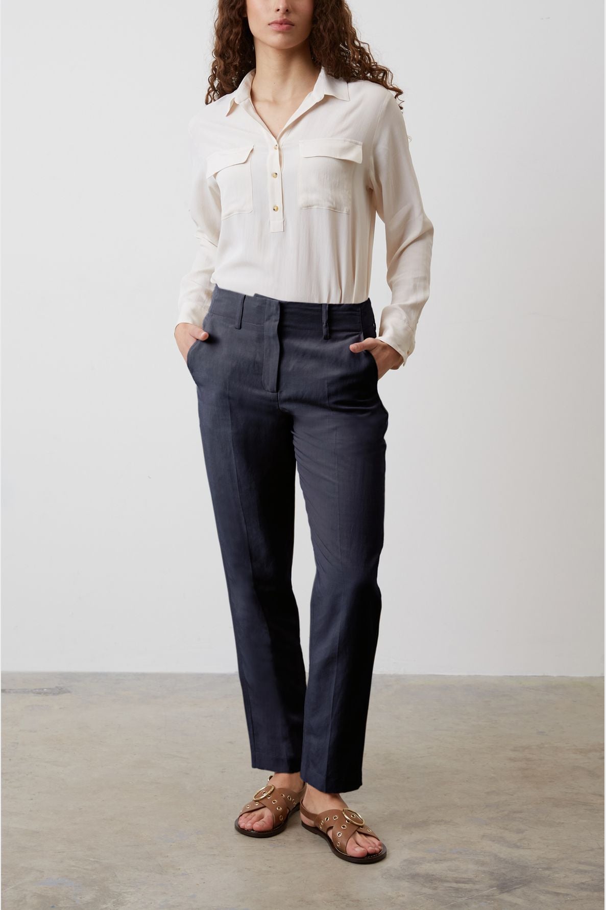 A person stands indoors wearing a cream blouse, dark tailored pants, and brown sandals. Their hands are in their pockets, and the background is plain with a concrete floor.