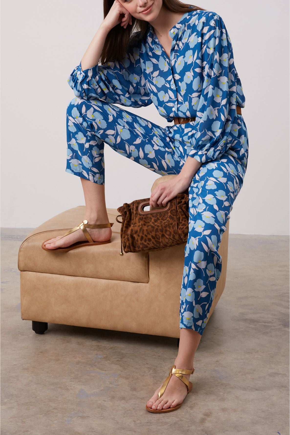 A person in a blue floral jumpsuit sits on a tan ottoman, holding a brown animal-print clutch. They wear gold sandals and have one arm resting on their knee.