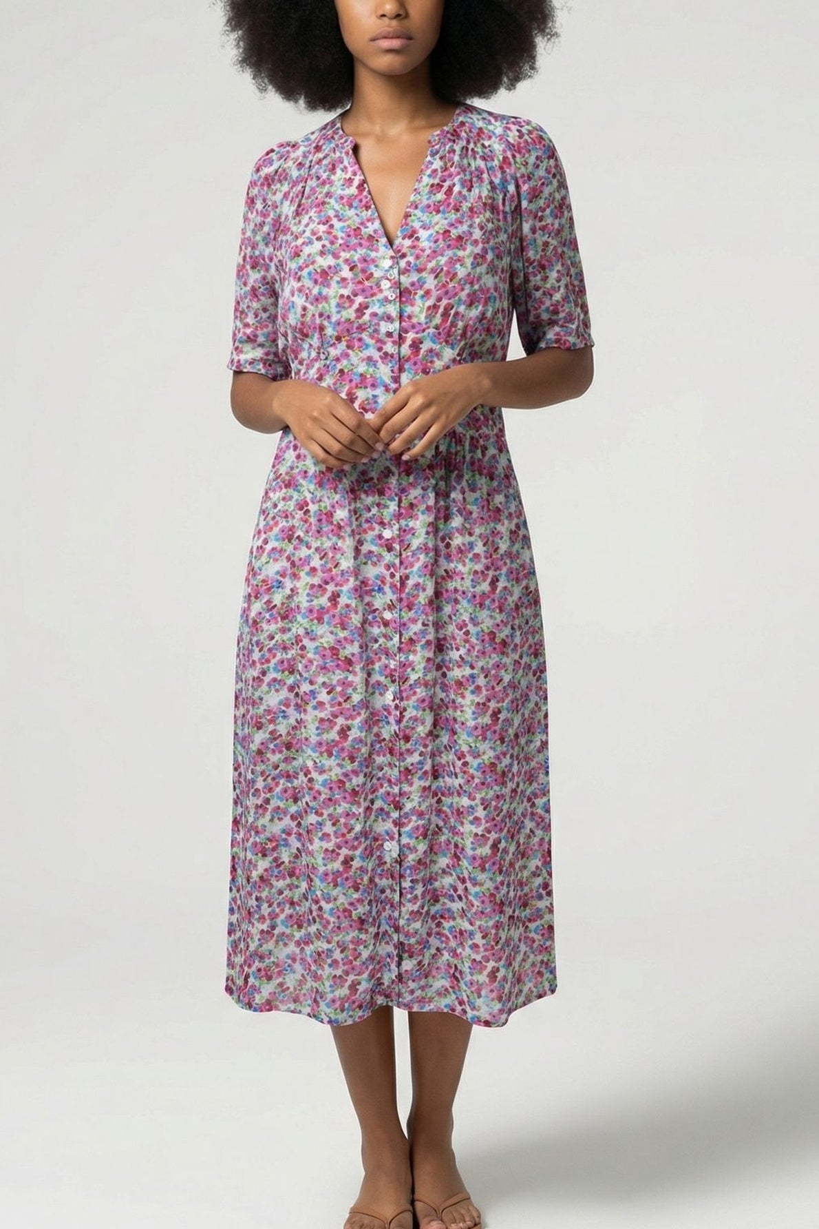 A woman wearing a robe mi-longue à motif floral, avec des touches de rose, de violet et de vert, se tient contre un fond clair et uni, ses mains délicatement jointes à sa taille. La matière toucher soie de la robe met en valeur l’élégance naturelle de sa silhouette.