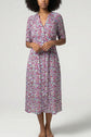 A woman wearing a robe mi-longue à motif floral, avec des touches de rose, de violet et de vert, se tient contre un fond clair et uni, ses mains délicatement jointes à sa taille. La matière toucher soie de la robe met en valeur l’élégance naturelle de sa silhouette.