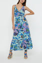 Woman wearing a sleeveless, V-neck, robe mi-longue with a blue, green, and purple imprimé floral pattern, standing with one hand on her hip and wearing wedge sandals.