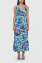 Woman wearing a sleeveless, ankle-length robe mi-longue with a bold blue, green, purple, and pink imprimé floral pattern, v-neckline, ruffled hem, and matching floral sandals. She stands against a plain light background.