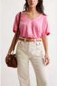 A woman wears a loose pink V-neck T-shirt tucked into high-waisted cream pants with a brown belt and carries a small leopard print bag over her shoulder.
