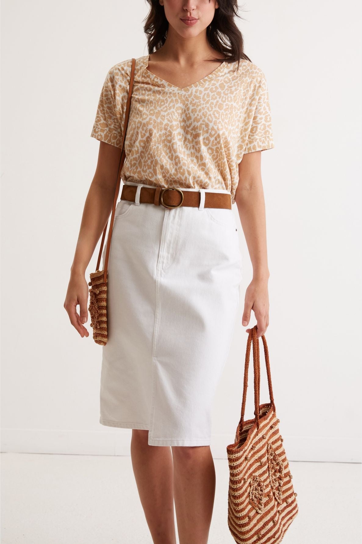 Woman wearing a beige animal print top, white knee-length skirt, brown belt, and carrying two brown woven bags, standing against a plain light background.