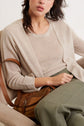 Woman sitting on a beige chair, wearing a light beige top, matching cardigan, olive green pants, and holding a brown leather bag on her lap. The photo is cropped at her shoulders and knees.