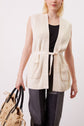 A person wearing a sleeveless cream knit gilet sans manche with a belt over a black top and dark jeans, holding a woven beige bag with black handles.