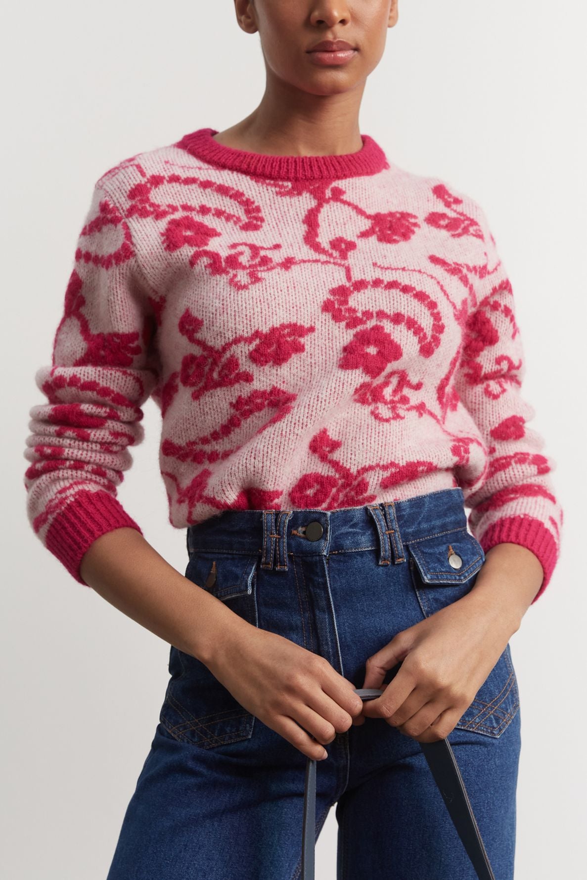 A person wearing a pink pull jacquard with red floral patterns and blue high-waisted jeans stands against a plain background, holding black straps in their hands.