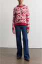 A person wearing a pink and red pull jacquard with blue flared jeans and brown shoes stands against a plain off-white wall and wooden floor.