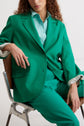 A person with long curly hair sits on a chair wearing a bright green suit and a light blue shirt, with one arm resting on the chair and the other on their leg.