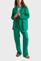 A person wearing a bright green suit with wide-legged trousers and a matching blazer over a light teal button-up shirt, paired with brown sandals, stands against a plain background.