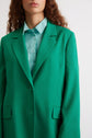 Woman wearing a bright green blazer over a light blue button-up shirt, standing against a plain light background. Her face is partially visible, and she has curly brown hair.