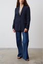 A person with long curly hair wears a dark blazer over a floral top, wide-leg blue jeans, and open-toe sandals, standing on a concrete floor against a plain white wall.