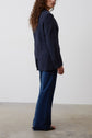 A person with curly hair stands sideways, wearing a navy blazer, dark blue wide-leg jeans, and open-toed sandals, against a plain light background with a concrete floor.