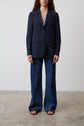 A person stands against a plain background wearing a dark blazer, printed shirt, wide-leg blue jeans, and black sandals.