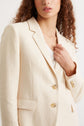 A woman with curly hair wears a cream-colored blazer with buttons and a white top underneath. She is holding the lapel of the blazer, shown from mid-face to waist.