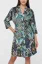 A woman wearing a knee-length robe with a colorful abstract pattern, featuring shades of blue, green, gold, and maroon, stands with one hand in the pocket and a slight smile. The design draws inspiration from a motif ethnique, adding an artisanal touch reminiscent of a veste kimono.