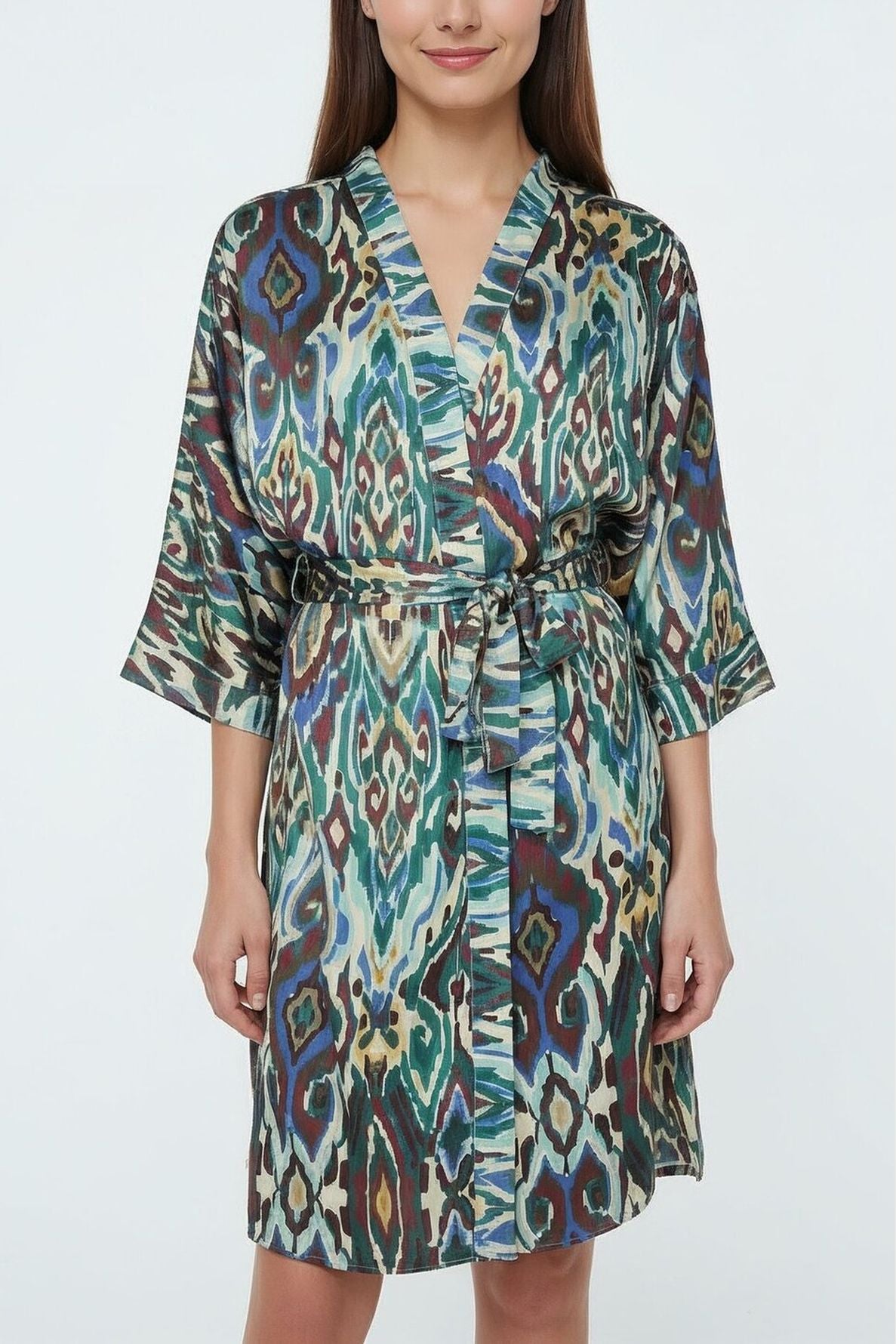 A woman wearing a colorful, patterned veste kimono with a ceinture amovible, standing against a plain white background. The robe features green, blue, brown, and cream abstract designs inspired by a motif ethnique.