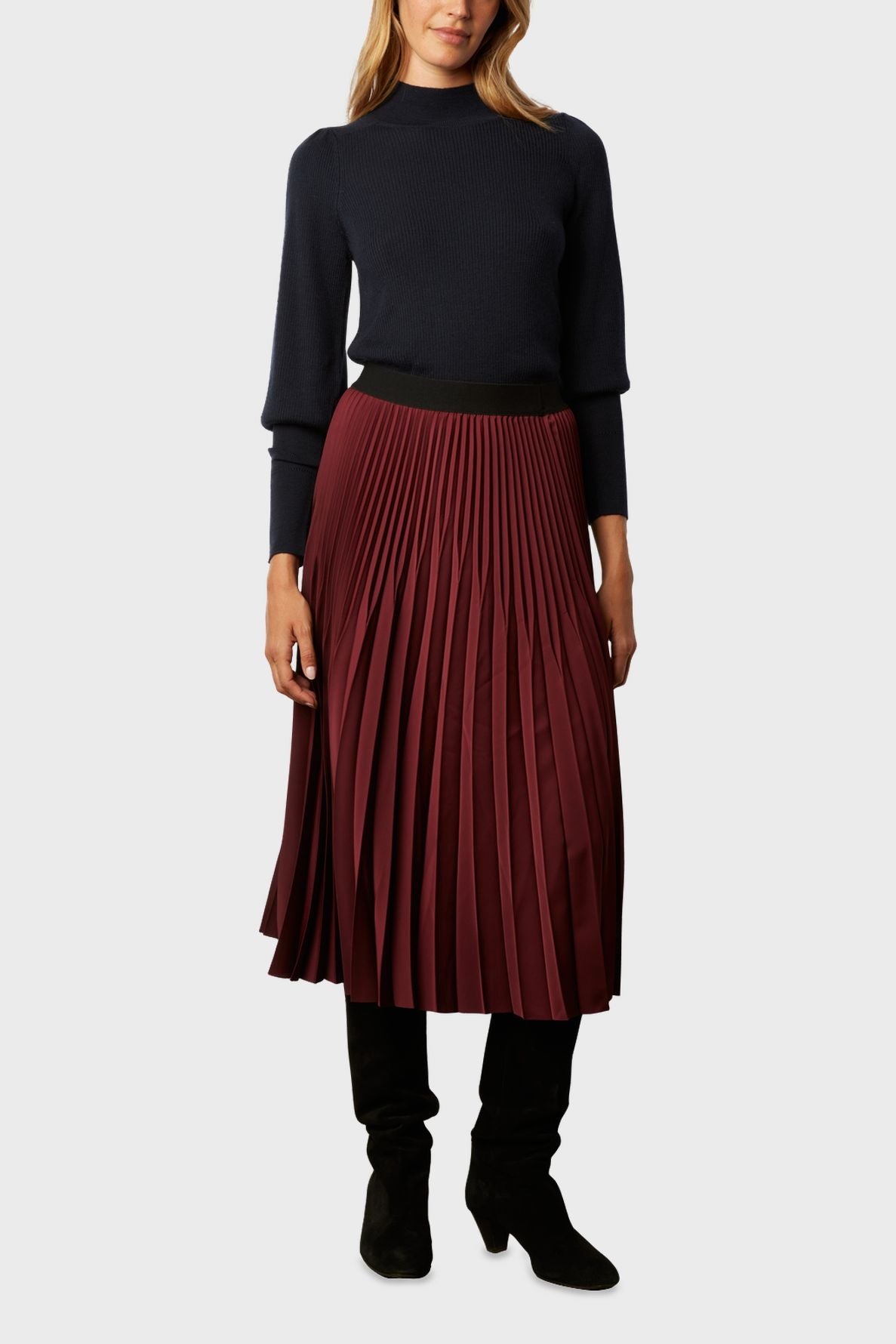 Woman wearing a navy long-sleeve sweater, a burgundy jupe midi plissée, and black knee-high boots, standing against a plain light gray background.