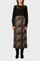 Woman wearing a jupe longue noire à motif floral rouge et beige, dotée d’une taille élastiquée, un pull noir à manches longues avec boutons dorés aux poignets, et des bottes noires, debout devant un fond uni.