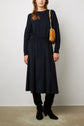 A woman stands against a plain background wearing a navy velours côtelé sweater, a matching navy jupe midi, black boots, and a tan shoulder bag with a gold chain strap for an effortlessly look casual-chic.