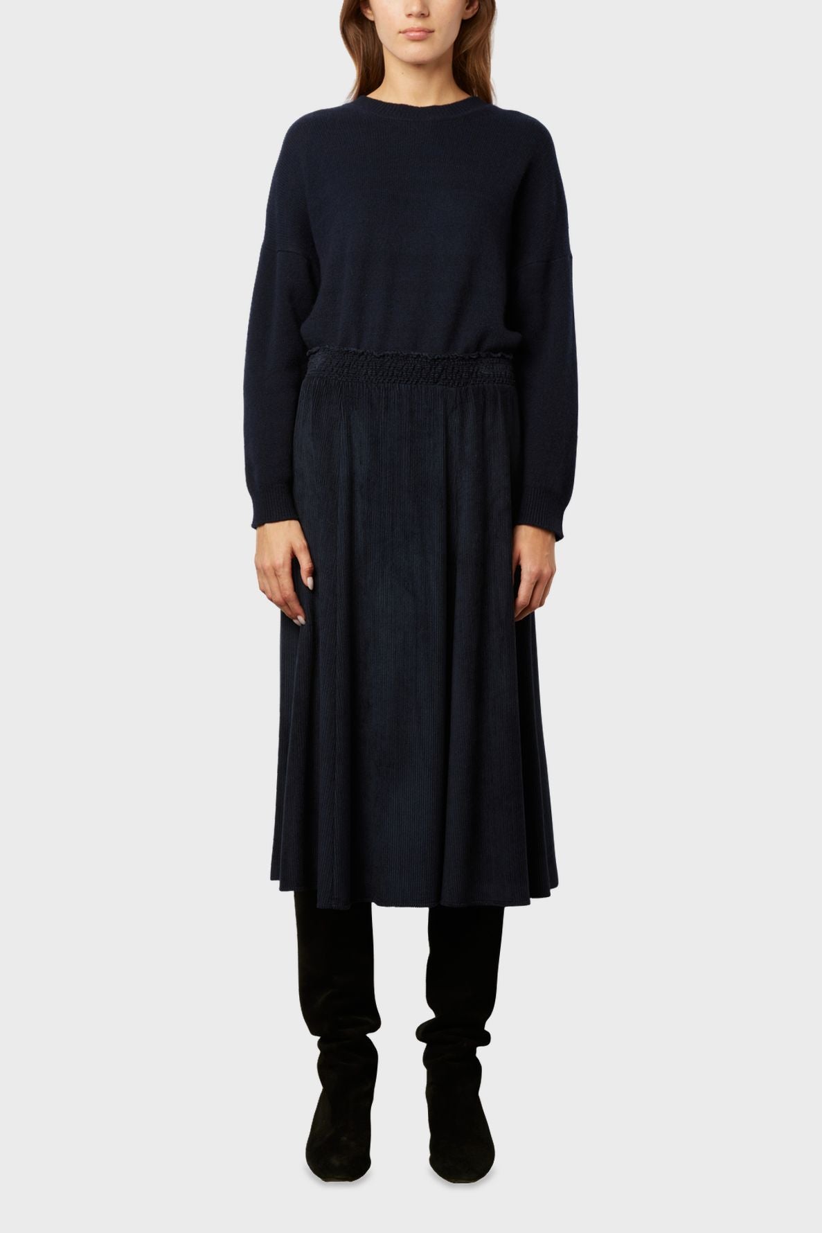 A woman stands facing forward wearing a navy blue sweater, a dark jupe midi plissée, and black boots against a plain light gray background for a look casual-chic.