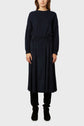 A woman stands facing forward wearing a navy blue sweater, a dark jupe midi plissée, and black boots against a plain light gray background for a look casual-chic.