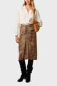 A woman wearing a cream blouse, a brown leopard print skirt, black knee-high boots, and carrying a tan shoulder bag stands against a plain light gray background.