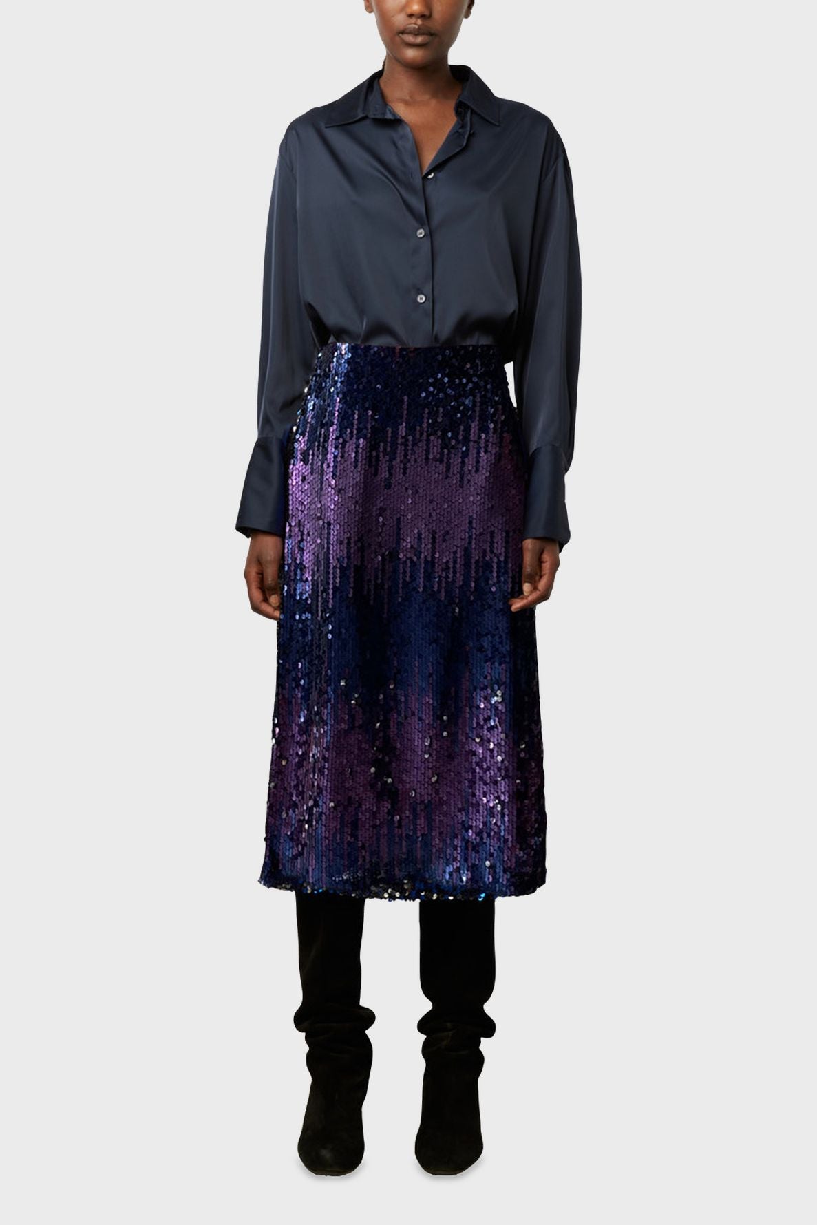 A person wearing a dark blue button-up shirt, a knee-length blue and purple sequin skirt, and black pants with black shoes, standing against a plain light gray background.