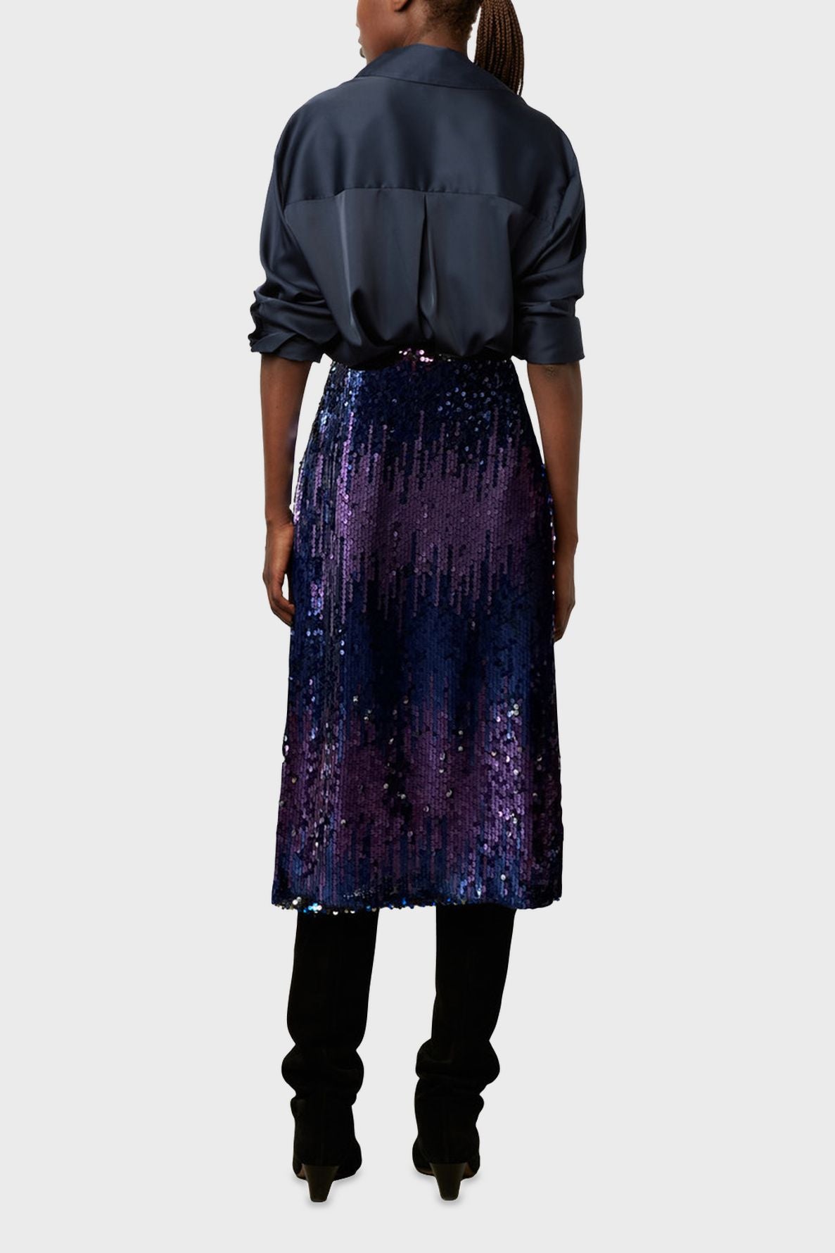 A person stands facing away, wearing a dark satin blouse, a shiny blue and purple sequin skirt, and black boots. The background is plain and light-colored.