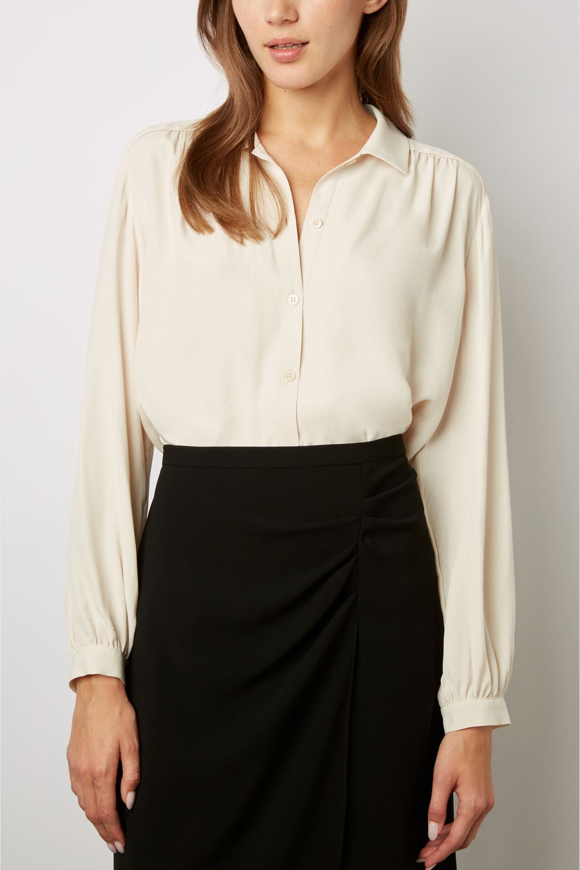 Woman wearing a long-sleeve, cream-colored blouse tucked into a high-waisted, black skirt with a subtle side pleat, standing against a plain background. Her face is partially out of frame.