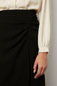 A person wearing a cream long-sleeve blouse tucked into a high-waisted black skirt with subtle draping, showing their right hand resting near the skirt.