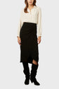 Woman wearing a long-sleeve cream blouse tucked into a black, knee-length wrap skirt with soft pleats, paired with tall black boots, standing against a plain light background.