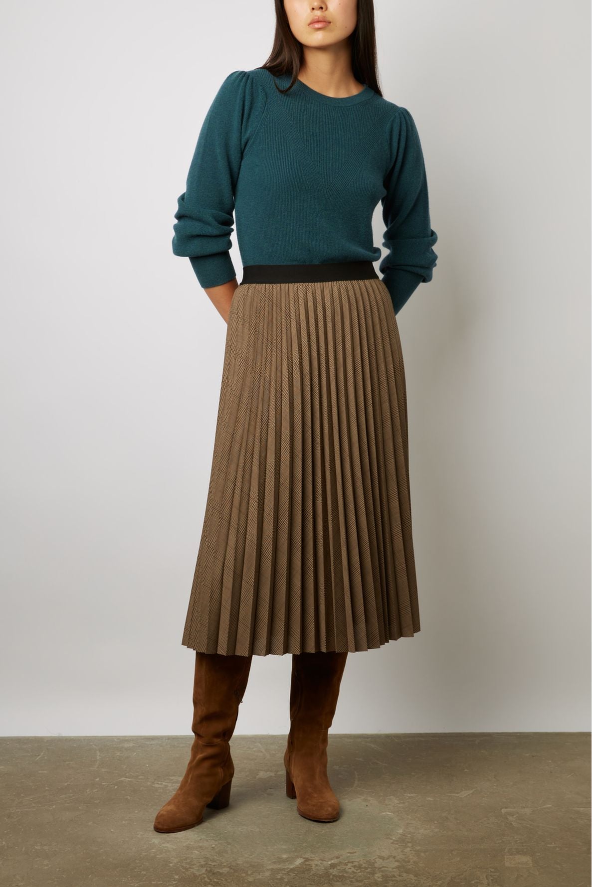 Person wearing a teal long-sleeve top, brown pleated midi skirt, and brown knee-high suede boots, standing against a plain light background.