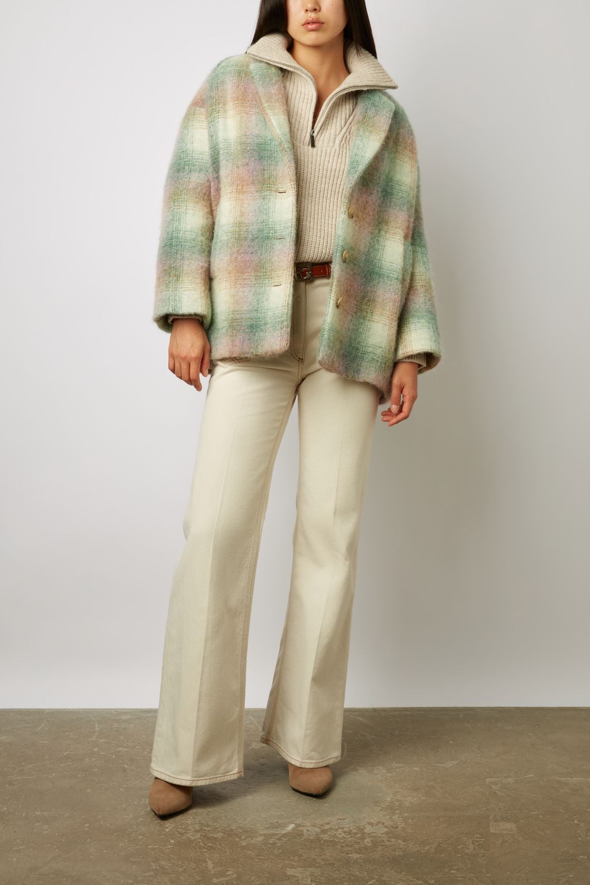 A woman wears a manteau court à carreaux in pastel tones crafted from laine mélangée over a beige zip-up sweater, cream wide-leg pants, and taupe heeled shoes, standing against a plain background.
