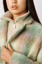 A woman wearing a beige knit sweater with a half-zip collar and a soft plaid manteau court in pastel green, beige, and pink tones, crafted from laine mélangée à carreaux, holding the coat closed with one hand. Only her lower face and upper torso are visible.