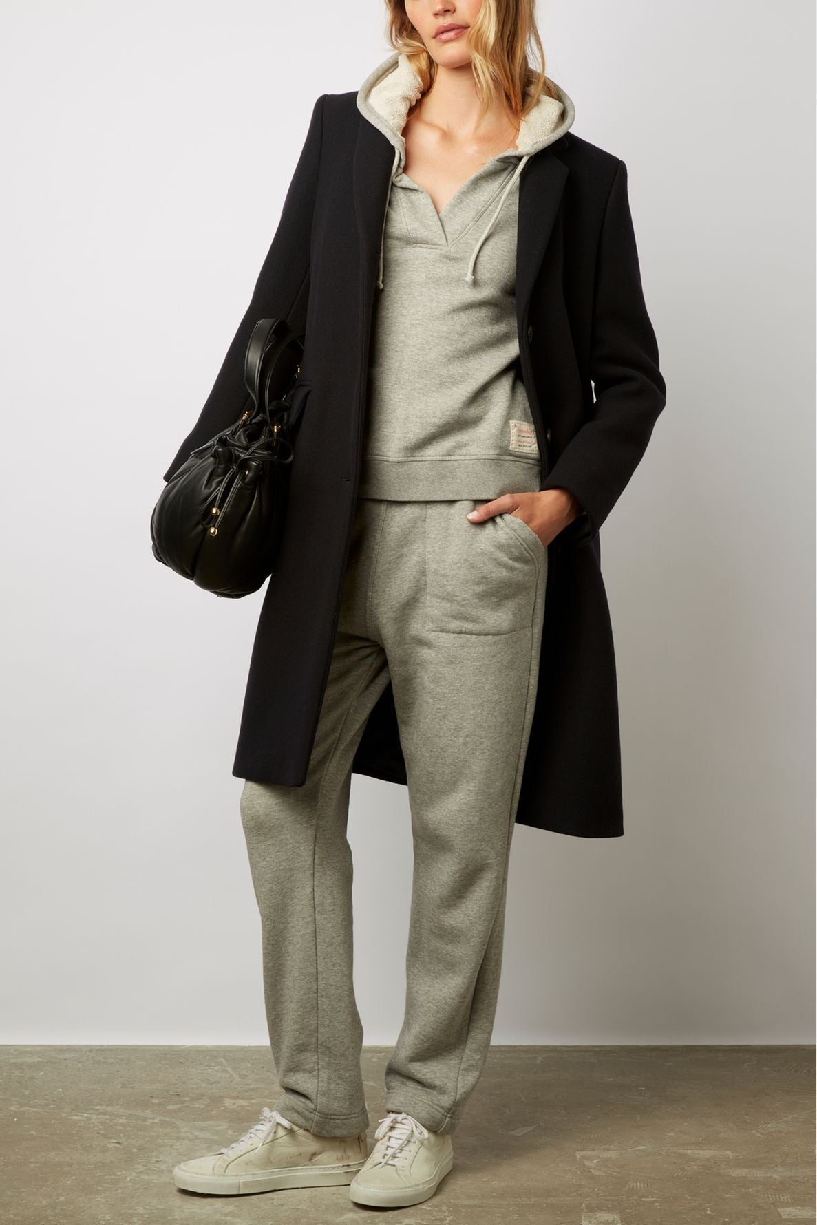 A person wearing a manteau droit mi-long in black wool, layered over a gray hoodie and matching sweatpants, with white sneakers, holds a black bag.