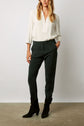 Woman wearing a white long-sleeve blouse, dark green pantalon cigarette, and black heeled boots, standing against a plain background with one hand in her pocket.