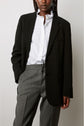 A person wearing a black blazer over a white button-up shirt and gray trousers stands with one hand in their pocket against a plain background.