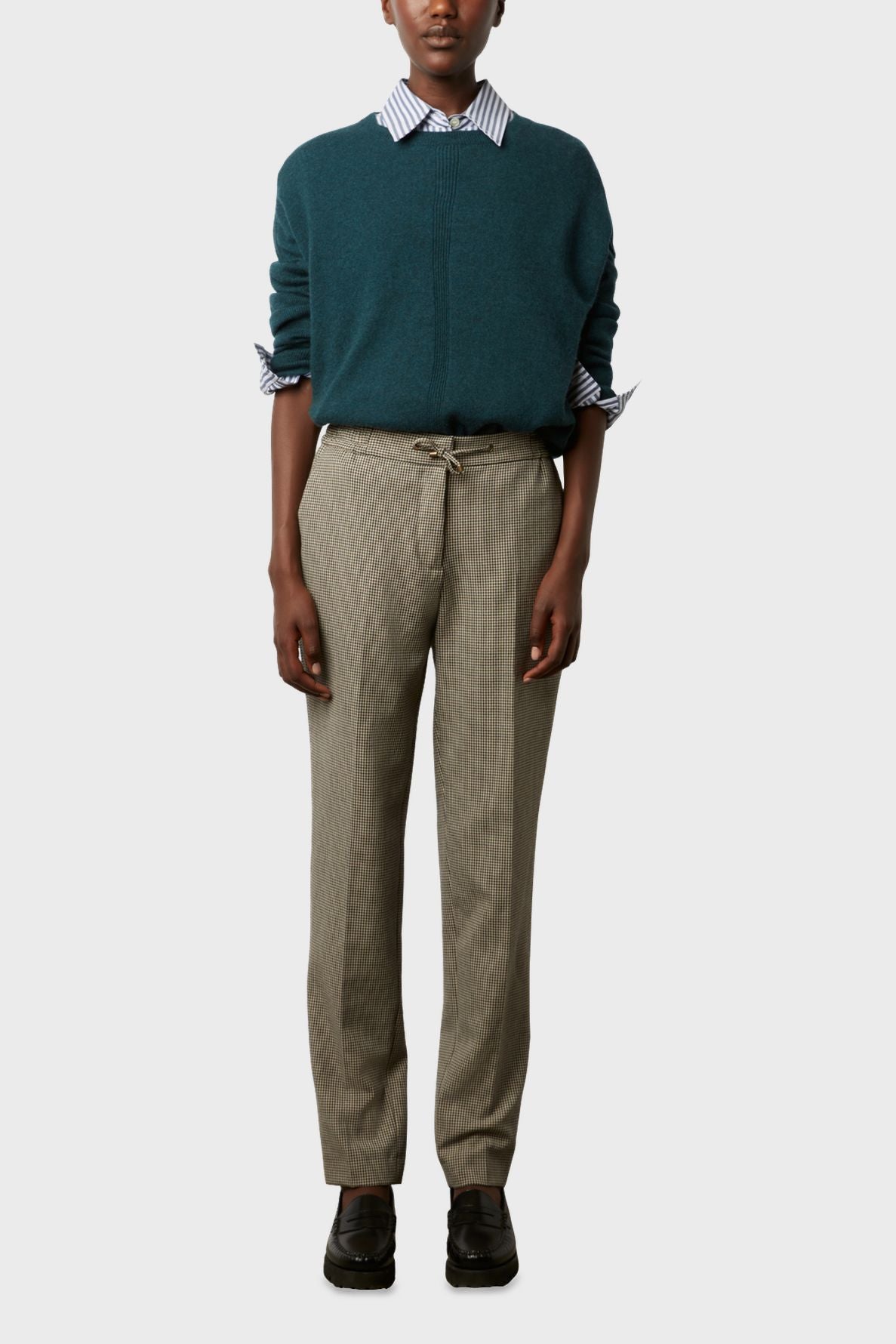 A person wears a teal sweater over a blue and white striped shirt, tan dress pants with a drawstring, and black shoes, standing against a plain light background.