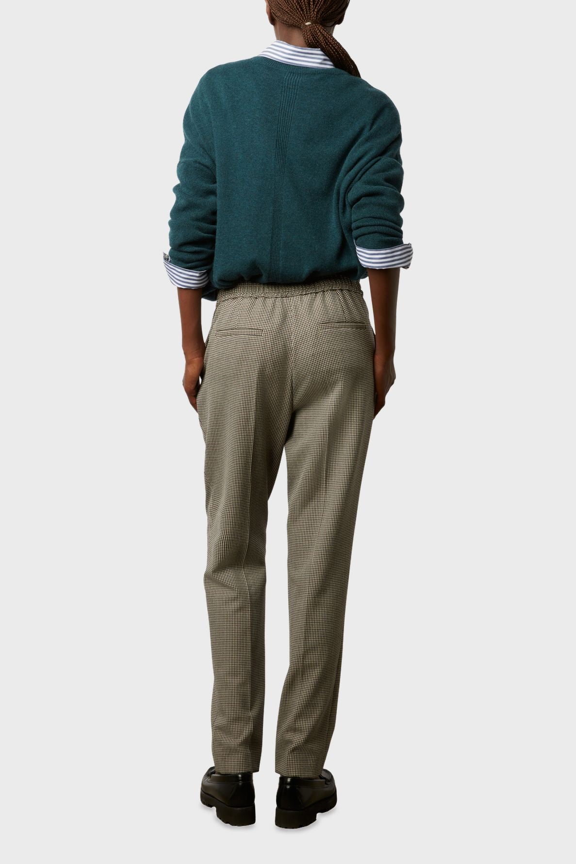 Person standing with back to camera, wearing a teal sweater over a striped shirt, beige trousers, and black shoes. The outfit looks casual and comfortable. The background is plain and light-colored.