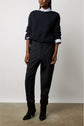 Person wearing a navy knit sweater over a white collared shirt, dark pantalon de tailleur en tissu uni, and black heeled boots stands against a plain background, with hands relaxed by their sides. Face is partially out of frame.